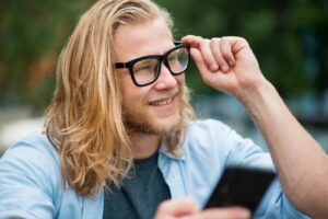 Jeune utilisateur souriant ajustant ses lunettes connectées tout en consultant son smartphone, illustration du débat sur la vie privée et la captation discrète par les smart glasses Meta Ray-Ban et alternatives Even Realities Brilliant Labs.