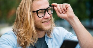 Jeune utilisateur souriant ajustant ses lunettes connectées tout en consultant son smartphone, illustration du débat sur la vie privée et la captation discrète par les smart glasses Meta Ray-Ban et alternatives Even Realities Brilliant Labs.