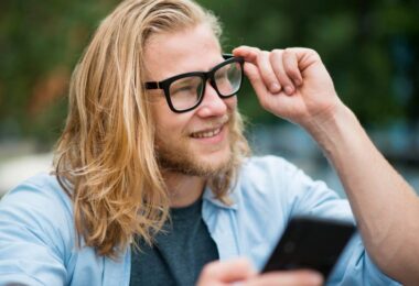 Jeune utilisateur souriant ajustant ses lunettes connectées tout en consultant son smartphone, illustration du débat sur la vie privée et la captation discrète par les smart glasses Meta Ray-Ban et alternatives Even Realities Brilliant Labs.