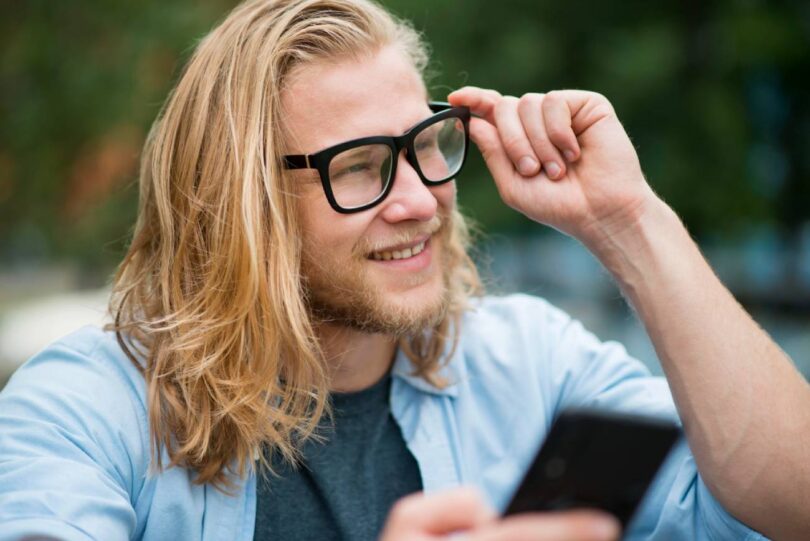 Jeune utilisateur souriant ajustant ses lunettes connectées tout en consultant son smartphone, illustration du débat sur la vie privée et la captation discrète par les smart glasses Meta Ray-Ban et alternatives Even Realities Brilliant Labs.