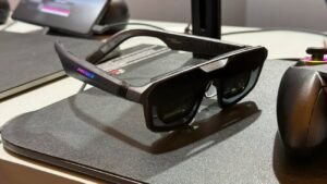 unettes AR gaming ASUS ROG Xreal R1 posées sur un tapis de souris noir avec LED RGB rose et bleu sur la branche, manette ROG en arrière-plan et station d'accueil Control Dock visible, premier modèle de lunettes display 240 Hz micro-OLED au monde.