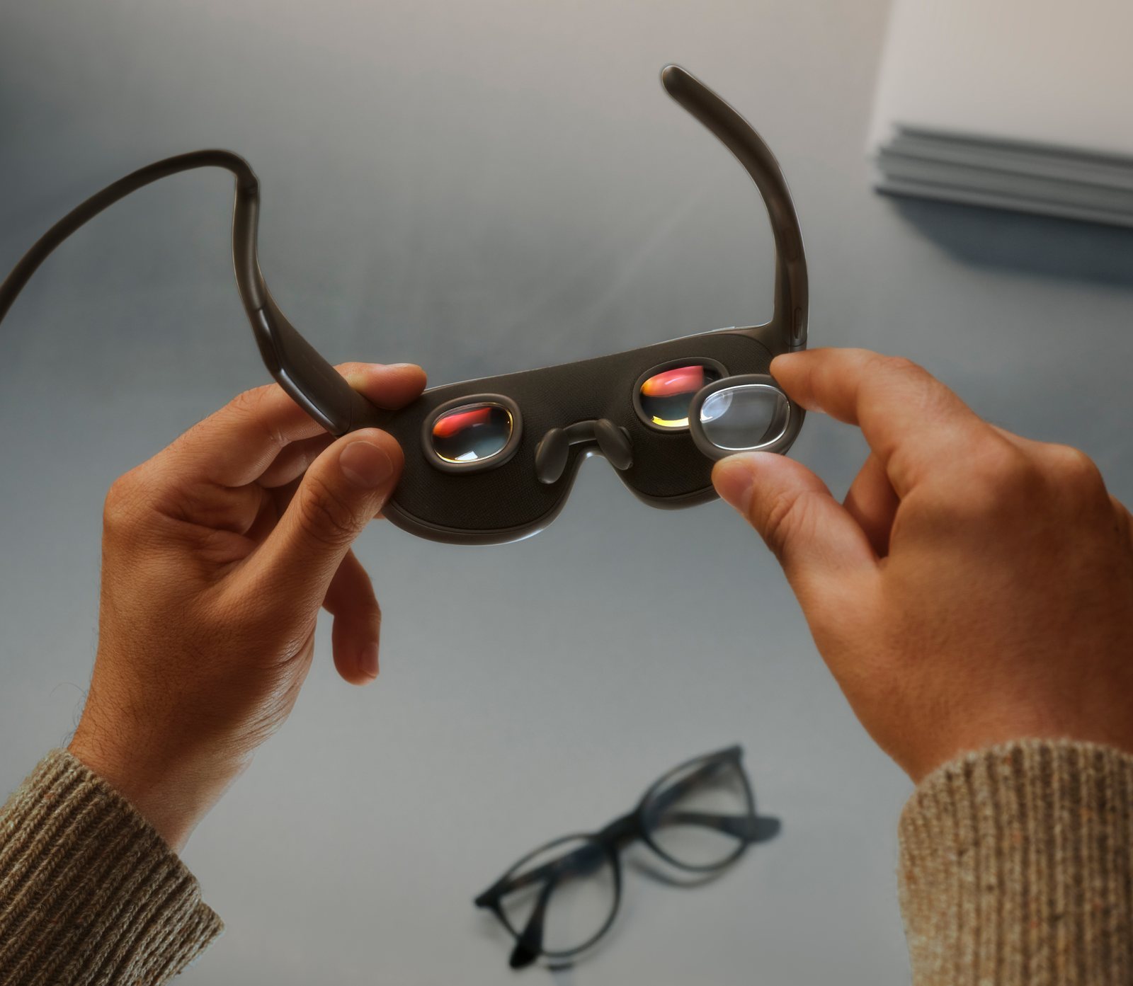 Arrière des Meta VR Glasses tenues à deux mains : deux oculaires, plaquettes de nez, câble relié à la branche et insert de verre correcteur en cours de pose, lunettes de vue classiques posées sur la table