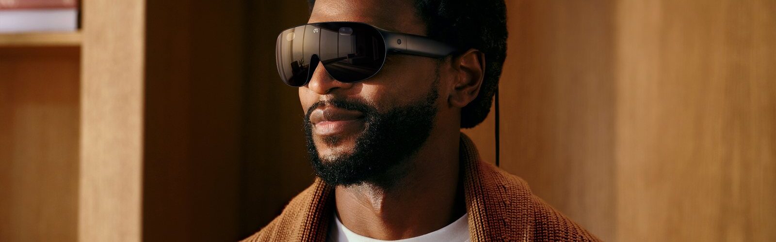 Meta VR Glasses portées par un homme en gilet en maille marron : lunettes de réalité virtuelle de 100 g à visière noire enveloppante, branches fines et câble optique relié au boîtier de calcul