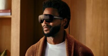 Meta VR Glasses portées par un homme en gilet en maille marron : lunettes de réalité virtuelle de 100 g à visière noire enveloppante, branches fines et câble optique relié au boîtier de calcul