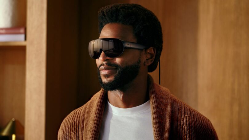 Meta VR Glasses portées par un homme en gilet en maille marron : lunettes de réalité virtuelle de 100 g à visière noire enveloppante, branches fines et câble optique relié au boîtier de calcul